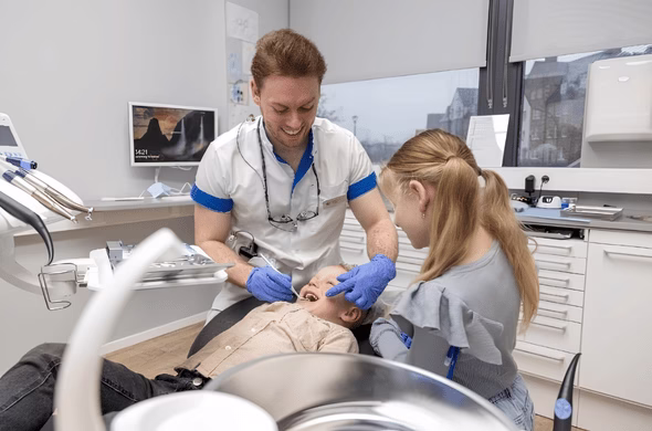 Tandarts behandelt een kind in de tandartsstoel terwijl een ander kind geïnteresseerd meekijkt.