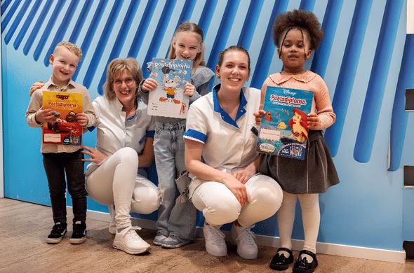 Twee kinderen poseren met twee medewerkers van een tandartspraktijk en houden tijdschriften en een kleurplaat vast.