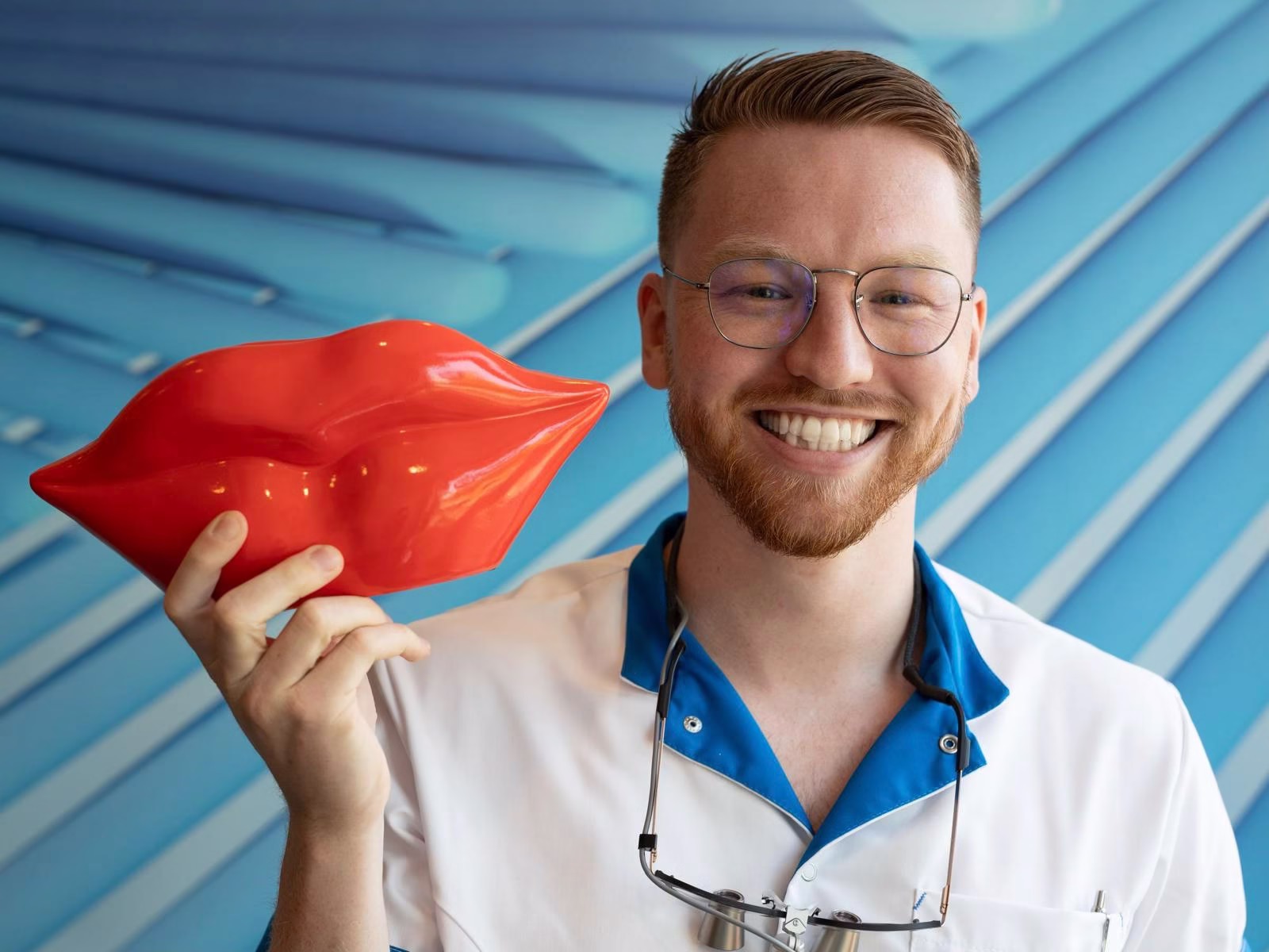 Lachende man met bril in een witte werkjas houdt een glanzend rood kunstobject in de vorm van lippen vast